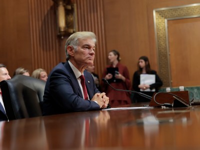 A person in a blue suit and red tie sits with his hands folded at a wooden table with a microphone in front of them. In the background is a row of people looking ahead in a large office room.
