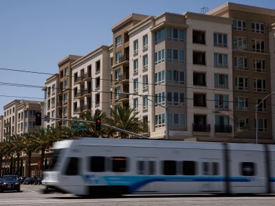 A white and blue train zooms by a street next two a multi-story apartment building in beige and cream tones.