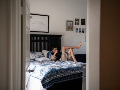 A young boy lounges on a bed with a blue and white patterned blanket, holding a camera, in a softly lit bedroom decorated with family photos and a framed sign that reads "It's always better when we're together."
