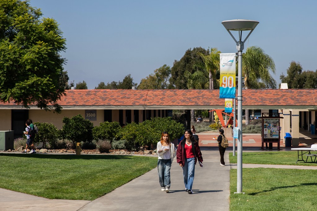 A sunny day on a community college campus where students walk along a concrete path flanked by green lawns and lush bushes. A building marked "3500" with a red-tiled roof is in the background. A banner hanging from a nearby lamppost celebrates the school's 90th anniversary.