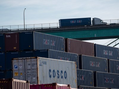 A stack of multicolored shipping containers, including brands like COSCO, Wan Hai, and tex, sits at the Port of Los Angeles. Above, a semi-truck carrying a blue container drives across a bridge, silhouetted against a light sky. The composition emphasizes the scale and flow of international freight transport.