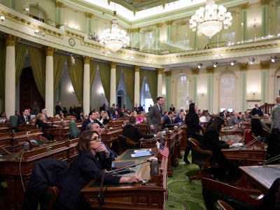 A legislative chamber filled with lawmakers seated at wooden desks. The room features high ceilings, large chandeliers, tall green curtains, and classical architectural details with white columns and gold trim.