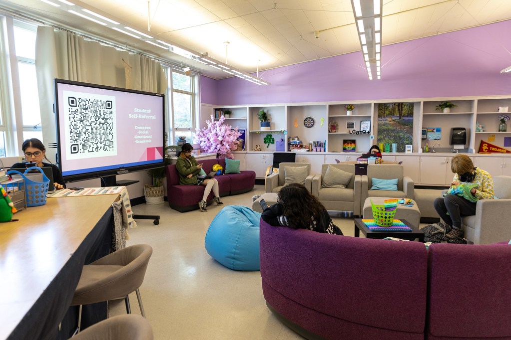 A wide view of a room with students sitting on couches or chairs as they stare down at their phones or laptops. The room is decorated in light purple tones and includes several activities and resources. A digital monitor displayed a slide with "student self-referral" information on it.