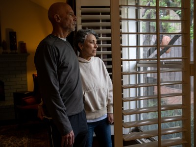 Two people hold each other as they stand in front of a window with its white shudders open, illuminating an outdoor area of a house.