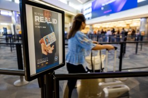 A sign posted on a line divider at an airport reminding travelers about getting their Real IDs before the deadline. A person walking through and pushing their luggage can be seen in the background.
