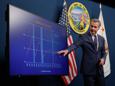 A person wearing a navy suit points to a monitor with a line chart on it during a press conference. The California state seal, state flag and the American flag can be seen behind them.